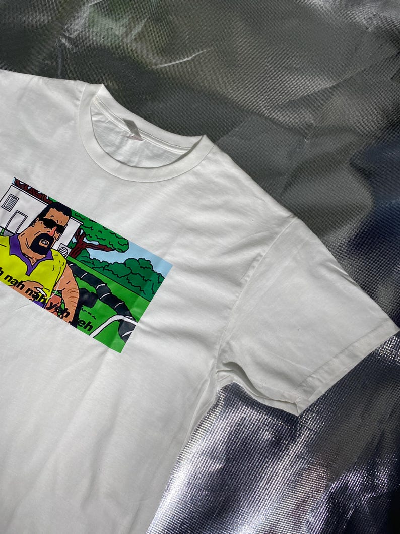 May include: White t-shirt featuring a cartoon image of a man in sunglasses and a yellow shirt. The image includes the text "Nah nah nah yeh eh". The shirt has a crew neck and short sleeves.