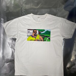 May include: White t-shirt featuring a cartoon image of a man in sunglasses and a yellow shirt, with the text "yeh nah nah yeh eh". The graphic is set against a green and blue background. The shirt is made of a soft, white material.