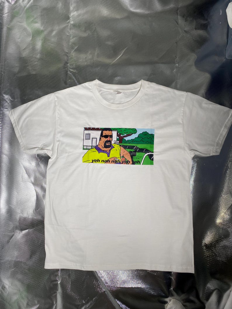 May include: White t-shirt featuring a cartoon image of a man in sunglasses and a yellow shirt, with the text "yeh nah nah yeh eh". The graphic is set against a green and blue background. The shirt is made of a soft, white material.