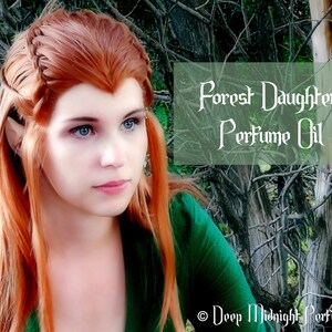 May include: A woman with long, red hair styled in a braid, wearing a green shirt, poses in front of a background of branches. The text "Forest Daughter Perfume Oil" is superimposed on the image.