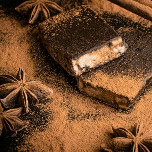 Op de afbeelding: Twee donkere chocolade repen met een lichtbruine vulling zijn bestoven met kaneelpoeder en omringd door steranijs peulen op een zwart oppervlak.