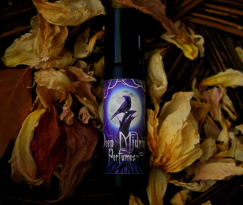 May include: A small, black roll-on bottle of perfume with a purple label featuring a raven perched on a branch under a full moon. The label reads "Deep Midnight Perfumes".