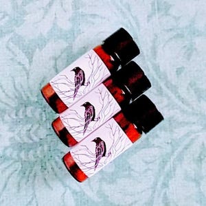 Op de afbeelding: Vier kleine glazen flesjes met zwarte doppen. Elk flesje heeft een etiket met een zwarte kraai op een tak. De etiketten hebben een witte achtergrond en een rode rand. De flesjes zijn gerangschikt op een lichtblauwe bloemenpatroon oppervlak.