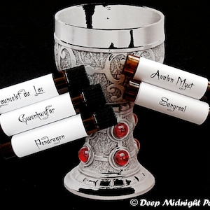 Könnte beinhalten: Fünf kleine Glasflaschen mit weißen Etiketten und schwarzer Beschriftung. Die Etiketten lauten "Lancelot du Lac", "Gwenhwyfar", "Pendragon", "Avalon Myst" und "Sangreal". Die Flaschen sind um einen silbernen Kelch mit roten Edelsteinen angeordnet.