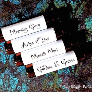 Op de afbeelding: Vijf kleine glazen flesjes met witte etiketten en zwarte letters. De etiketten lezen "Mourning Glory", "Ashes of Love", "Memento Mori", "Gardens & Graves" en "Deep Midnight Perfumes".