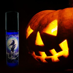 May include: A blue glass roll-on bottle with a label featuring a black raven perched on a branch in front of a full moon. The label also includes the text "Deep Midnight Perfumes". The bottle is in front of a lit jack-o'-lantern.