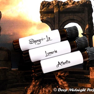 Op de afbeelding: Drie kleine bruine glazen flesjes met witte etiketten en zwarte dopjes. De etiketten zijn op elkaar gestapeld en hebben de namen "Shangri-La", "Lemuria" en "Atlantis" in zwarte cursieve letters. De flesjes staan voor een wazige achtergrond van een stenen boog en een standbeeld.