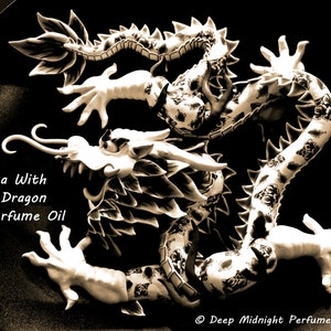 May include: A black and white ceramic dragon figurine with intricate details. The dragon is curled up with its head raised and its claws extended. The text "Tea With A Dragon Perfume Oil" is written below the dragon.