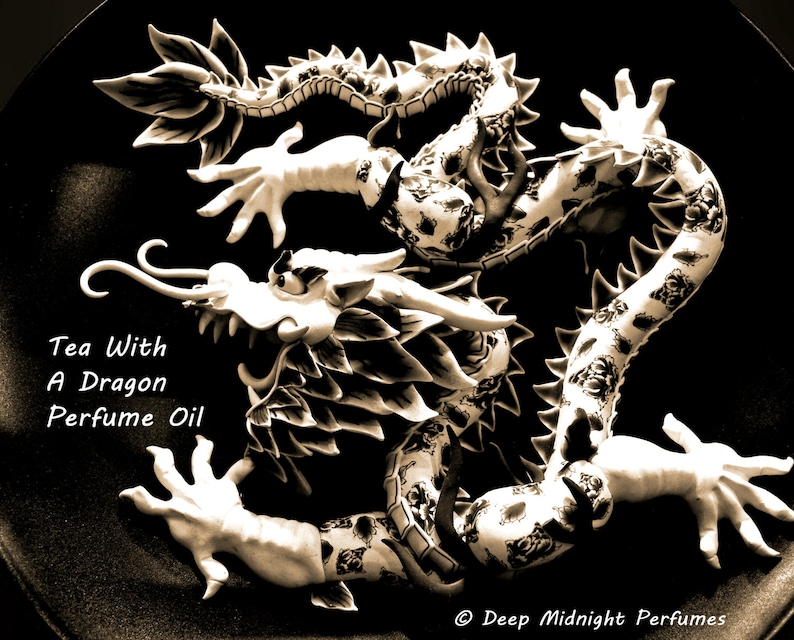 May include: A black and white ceramic dragon figurine with intricate details. The dragon is curled up with its head raised and its claws extended. The text "Tea With A Dragon Perfume Oil" is written below the dragon.