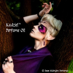 May include: A person with purple and silver eye makeup and pointed ears is wearing a purple shawl and a silver necklace with a crystal pendant. The text "Kadziel™ Perfume Oil" is visible in the image.