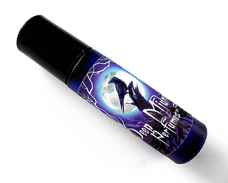 Op de afbeelding: Een donkerblauwe glazen fles met een zwarte dop en een etiket met de tekst "Deep Midnight Perfumes" met een raaf en een maanmotief.