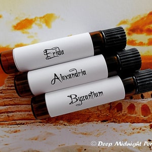 Może przedstawiać: Trzy małe szklane butelki z czarnymi korkami i białymi etykietami. Etykiety mają nazwy "Fridu", "Alexandria" i "Byzantium" napisane czarnym pismem odręcznym. Butelki są ułożone w kształcie piramidy na piaszczystej powierzchni.