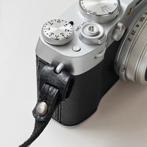 May include: Close-up of a silver digital camera with black leather strap. The camera has multiple dials and buttons, including a dial with numbers and a power switch. The strap is attached with a metal clasp and a silver button.