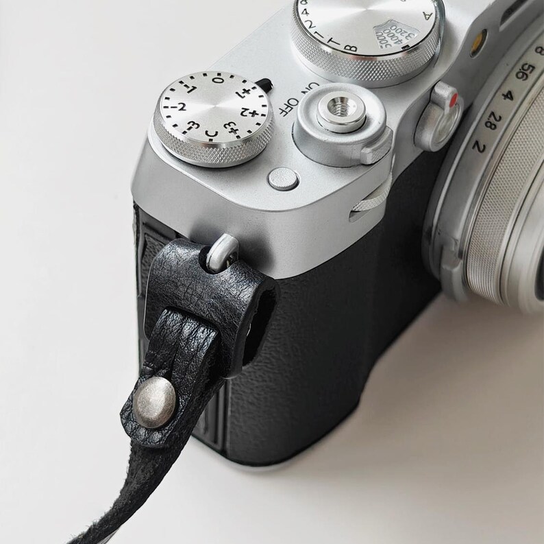 May include: Close-up of a silver digital camera with black leather strap. The camera has multiple dials and buttons, including a dial with numbers and a power switch. The strap is attached with a metal clasp and a silver button.