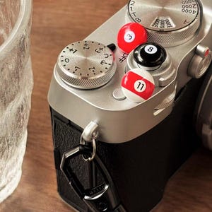 May include: A silver camera with three billiard ball-shaped accessories. One is red with a white 3, another is black with a white 8, and the third is white with red stripes and a white 11. The camera has dials and a black leather grip.