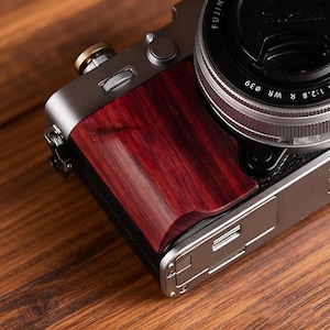 Könnte beinhalten: Nahaufnahme einer silber-schwarzen Kamera mit einem dunkelroten Holzgriff. Das Objektiv ist schwarz mit silbernen Details. Die Kamera liegt auf einer Holzoberfläche.