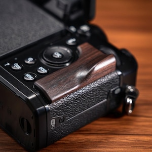 Könnte beinhalten: Nahaufnahme einer schwarzen Digitalkamera mit einem Griff aus dunklem Holz. Der Griff ist tiefbraun und hat ein geschwungenes Design. Das Kameragehäuse hat Knöpfe und eine strukturierte Oberfläche.