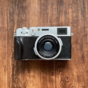 Op de afbeelding: Een zilveren en zwarte digitale camera met een zwarte lens en een houten handgreep. De lens is gemarkeerd met "Fujinon Aspherical Lens" en "Super EBC f=23mm 1:2 E". De camera ligt op een houten oppervlak.