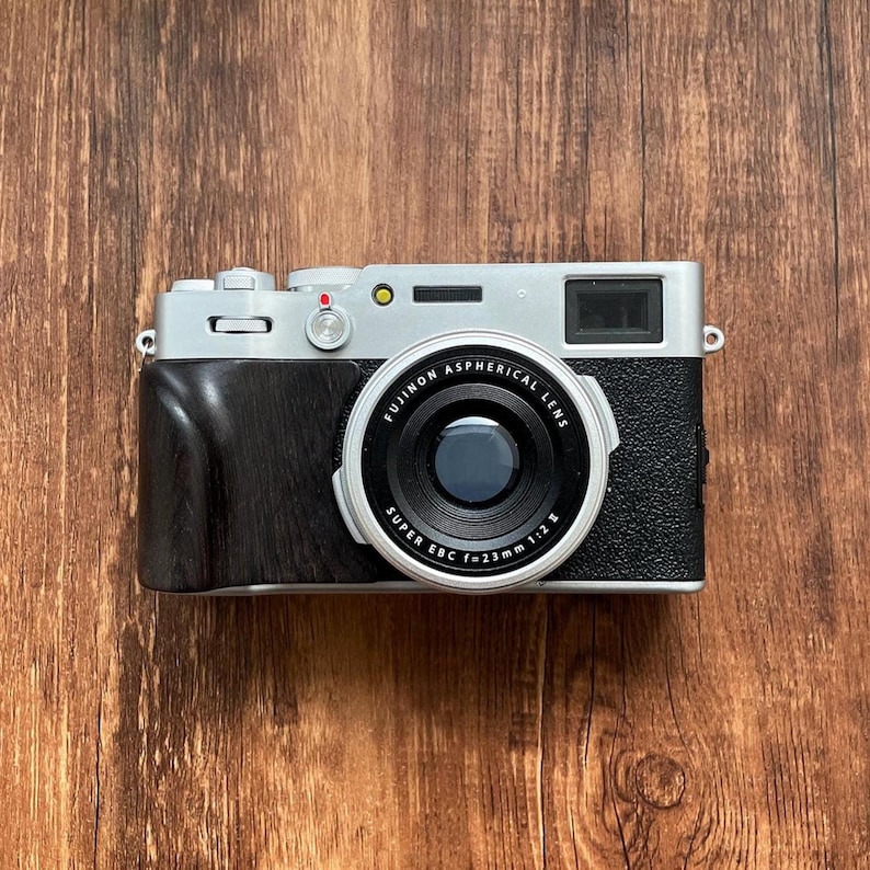 Op de afbeelding: Een zilveren en zwarte digitale camera met een zwarte lens en een houten handgreep. De lens is gemarkeerd met "Fujinon Aspherical Lens" en "Super EBC f=23mm 1:2 E". De camera ligt op een houten oppervlak.