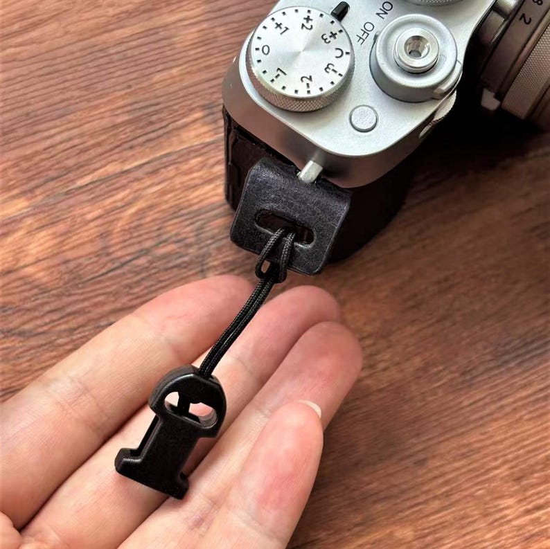 May include: A black camera wrist strap attached to a silver camera. The strap features a small, rectangular leather piece and a black cord that connects to a plastic clasp. The camera has dials and buttons.