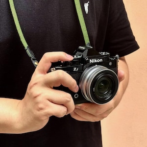 May include: Black Nikon Z f camera with a silver lens, held by a person. The camera has a green strap around the neck. The person is wearing a black shirt with a white logo.