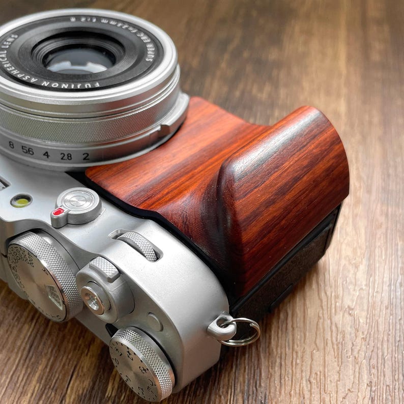 Op de afbeelding: Zilveren digitale camera met een roodbruine houten handgreep. De camera heeft een zilveren lens en diverse draaiknoppen en knoppen. De houten handgreep vertoont een duidelijk houtnerfpatroon.