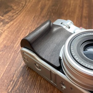 Op de afbeelding: Close-up van een zilveren camera met een zwarte houten grip. De lens is zilver en zwart met de tekst "SUPER EBC f=23 mm". De camera staat op een houten oppervlak.