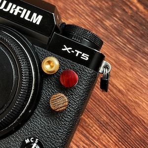 May include: Close-up of a black Fujifilm X-T5 camera with three decorative buttons in gold, red, and wood grain. The camera body has a textured leather-like finish, and the lens is visible on the left side. The background is a wooden surface.