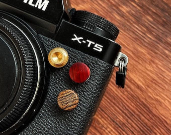 May include: Close-up of a black Fujifilm X-T5 camera with three decorative buttons in gold, red, and wood grain. The camera body has a textured leather-like finish, and the lens is visible on the left side. The background is a wooden surface.