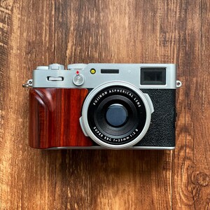 Op de afbeelding: Een zilveren en zwarte digitale camera met een houten handgreep en een zwarte lens. De lens is voorzien van de tekst "FUJINON ASPHERICAL LENS" en "SUPER EBC f=23mm 1:2 #". De camera staat op een houten ondergrond.