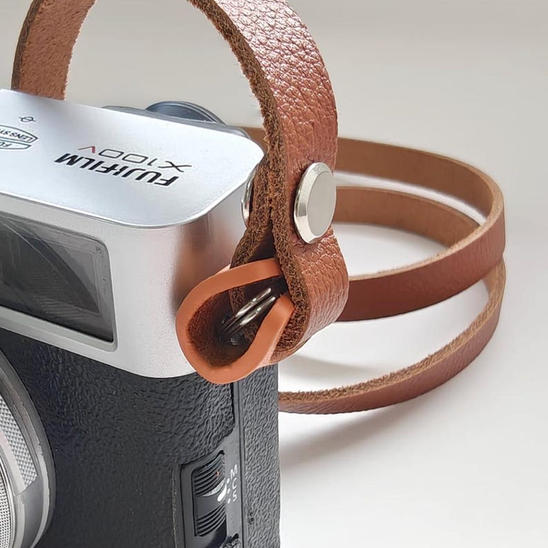 May include: Close-up of a Fujifilm X100V camera with a brown leather wrist strap. The camera is silver and black. The strap is attached with a silver rivet and an orange leather loop.