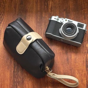 May include: A dark charcoal gray rectangular pouch with a beige strap closure featuring a silver snap button and a matching beige wrist strap attached to a gold-toned metal clasp, positioned next to a silver and black digital camera.
