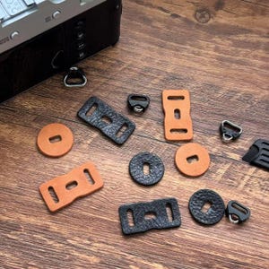 May include: A collection of camera accessories, including leather strap connectors and metal rings, in black and tan. The items are arranged on a wooden surface, with a black camera body visible in the background.