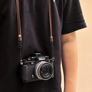 May include: Black Nikon Zf camera with a silver lens, suspended by a brown leather strap. The camera body is black, and the lens is silver. The strap is attached to the camera with small metal clasps.
