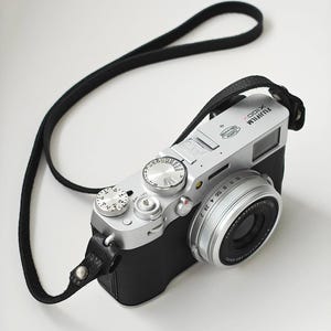 May include: A silver and black Fujifilm X100V digital camera with a black leather strap. The camera has multiple dials and a lens with a silver ring. The camera is on a white surface.