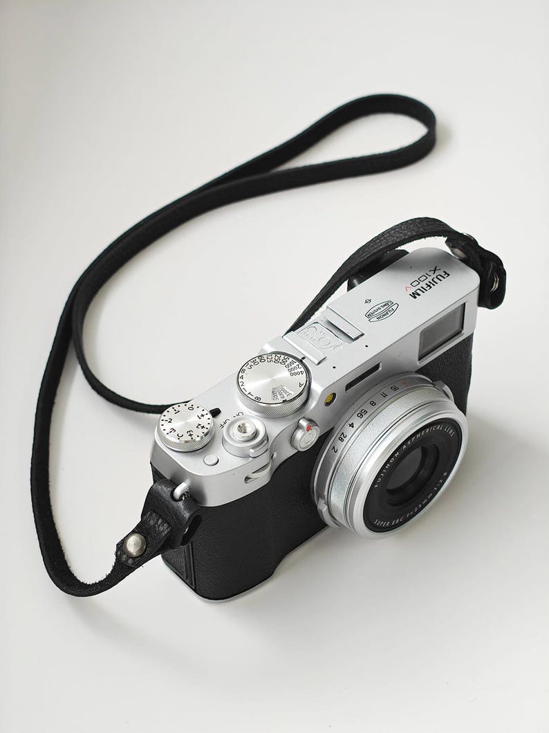 May include: A silver and black Fujifilm X100V digital camera with a black leather strap. The camera has multiple dials and a lens with a silver ring. The camera is on a white surface.