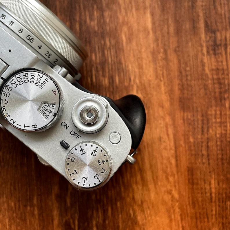 Op de afbeelding: Zilveren digitale camera met zwarte grip, met draaiknoppen voor sluitertijd, diafragma en belichtingscompensatie. De lens heeft markeringen voor brandpuntsafstanden. De camera staat tegen een warme, houten achtergrond.