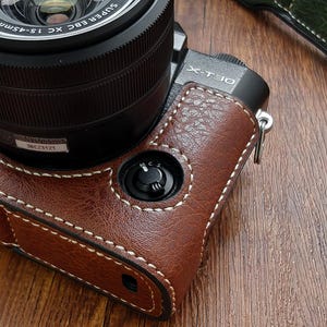 May include: A brown leather camera case with white stitching, fitted to a black camera body. The camera lens is black with silver text. A green leather case is visible in the background.
