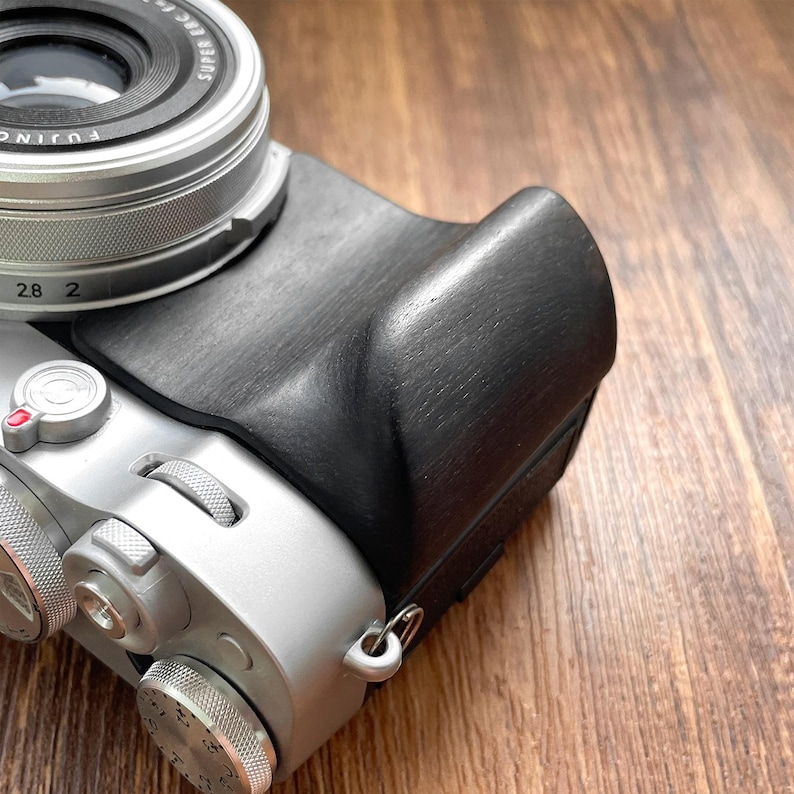 Op de afbeelding: Een zwarte leren cameragrip bevestigd aan een zilveren camerabehuizing. De grip is getextureerd en gevormd om de vorm van de camera te passen. De cameralens is zichtbaar, met een zilveren ring en zwarte details. De achtergrond is een houten oppervlak.