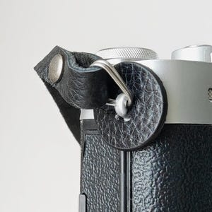 Op de afbeelding: Close-up van een zwarte leren camerariem bevestigd aan een zilveren camerabehuizing. De riem is bevestigd met een metalen klinknagel en een zilveren metalen ring. De camerabehuizing heeft een getextureerde, zwarte lederen afwerking.