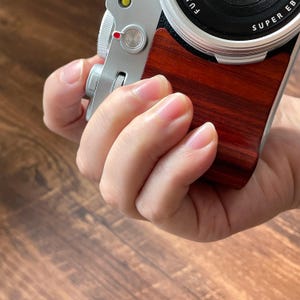Op de afbeelding: Een zilveren camera met een donkerhouten handvat wordt in een hand gehouden. De lens is zwart met zilveren rand en de woorden "SUPER EB" zijn zichtbaar. De houten handgreep heeft een rijke, roodbruine kleur.