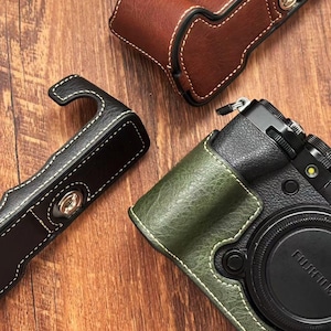 May include: Leather camera cases in brown, black, and green, with white stitching. The cases are designed to fit a camera, with a Fujifilm lens visible. The cases have a textured appearance and metal hardware.