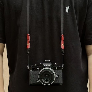 Puede incluir: Cámara Nikon Z f negra con lente negra y correa roja y negra. La cámara cuelga de una correa negra con detalles de cuero rojo. La correa de la cámara está sujeta a la cámara.