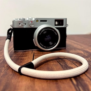 May include: A black and silver digital camera with a white wrist strap. The camera has a silver lens and a black body. The wrist strap is made of a woven white material with black accents.