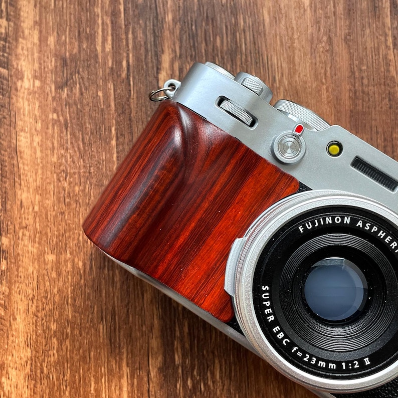 Op de afbeelding: Een zilveren en houtnerf digitale camera met een zwarte lens. De camera heeft een rode en gele knop en de lens leest "FUJINON ASPHERICAL SUPER EBC f=23mm 1:2 II". De houtnerf grip is roodbruin van kleur.