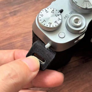 May include: Close-up of a silver camera with black leather strap attachment. The camera has dials and buttons, including an exposure compensation dial. A black leather strap is being attached to the camera.