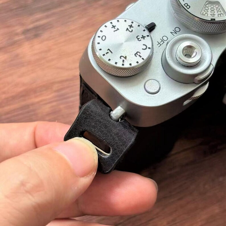 May include: Close-up of a silver camera with black leather strap attachment. The camera has dials and buttons, including an exposure compensation dial. A black leather strap is being attached to the camera.