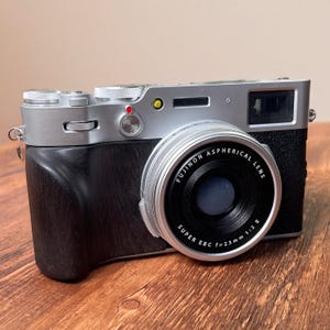 Op de afbeelding: Een zilveren en zwarte Fujifilm X100V digitale camera met een 23mm f/2 lens. De camera heeft een zwarte lederen grip en zilveren draaiknoppen. De lens is gemarkeerd met "Fujinon Aspherical Lens". Op een houten oppervlak.