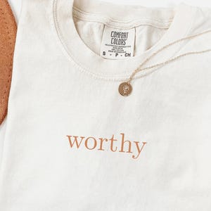 Puede incluir: Camiseta color crema con la palabra "worthy" en escritura color cobre. Un collar dorado con un colgante descansa sobre la camiseta. Un sombrero marrón y hojas de palma secas también están en la imagen.