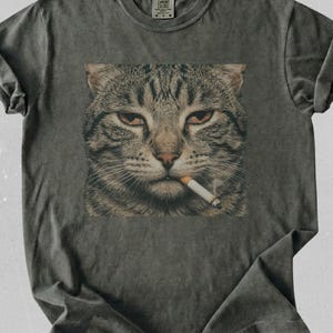Puede incluir: Una camiseta gris carbón de manga corta con un estampado gráfico cuadrado de un gato atigrado con un cigarrillo en la boca. La camiseta tiene cuello redondo y un acabado lavado vintage. La etiqueta de la marca Comfort Colors es visible en el cuello.